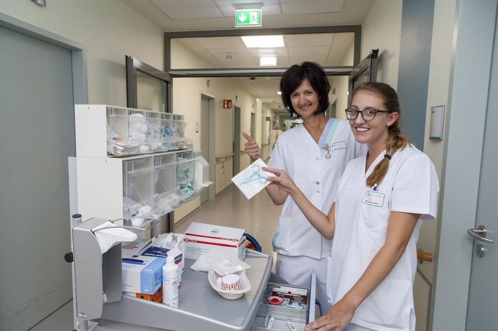 Das Foto zeigt zwei Pflegefachkräfte dem Utensilienwagen zur Blutabnahme unterwgs auf dem Krankenhausflur 