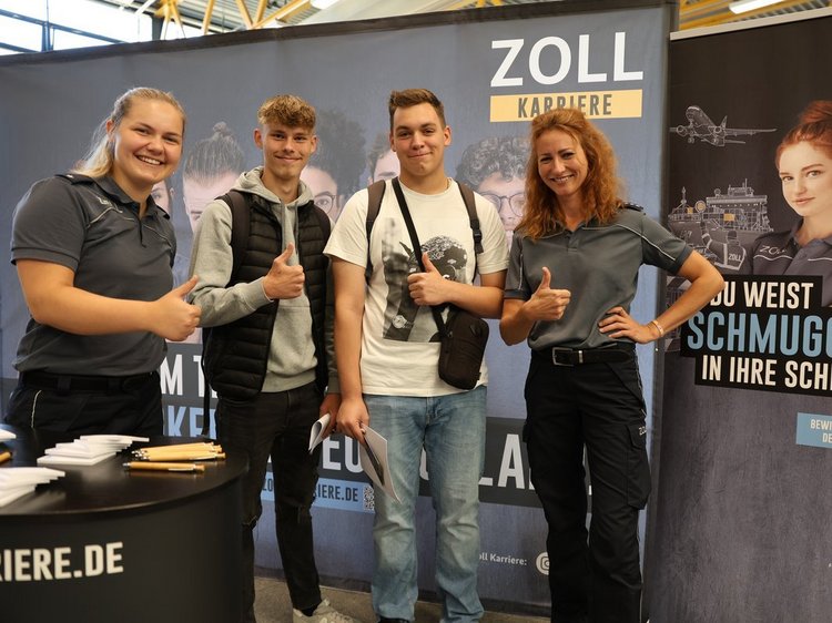 Das Foto zeigt 4 Personen vor dem Messestand des Zolls, die in die Kamera lachen