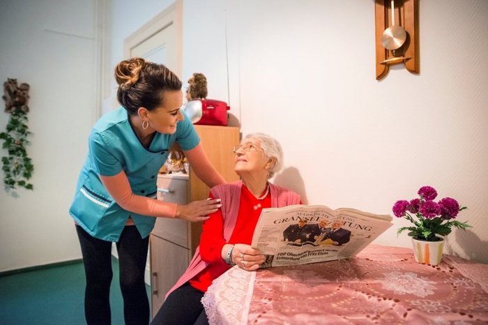 Das Fotozeigt eine Seniorin mit Zeitung und eine Pflegerin, die sie freundlich anspricht